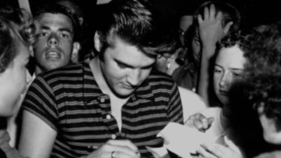 Elvis Presley fashion icon wearing a T-shirt - signing an autograph with loads of fans around him